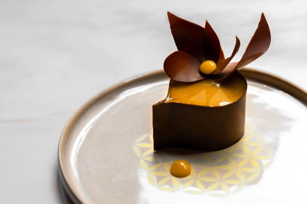 Dessert raffiné en forme de demi-cœur, enrobé d'un glaçage caramel brillant et entouré d'une fine coque en chocolat. Décoré d'une élégante fleur en chocolat et d'une touche de crème dorée, le tout présenté sur une assiette en céramique au design géométrique